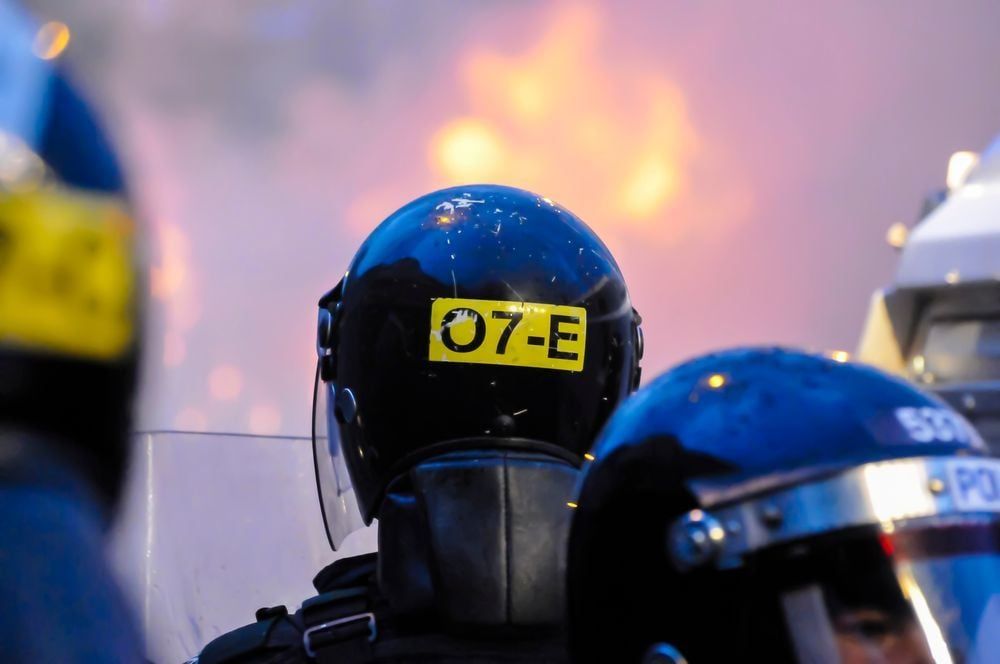 People wear riot gear with riot shields. In the background is fire. 