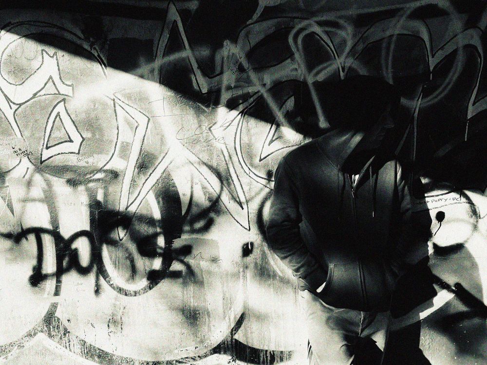 Guy in a hoodie, leaning against a graffitied wall.