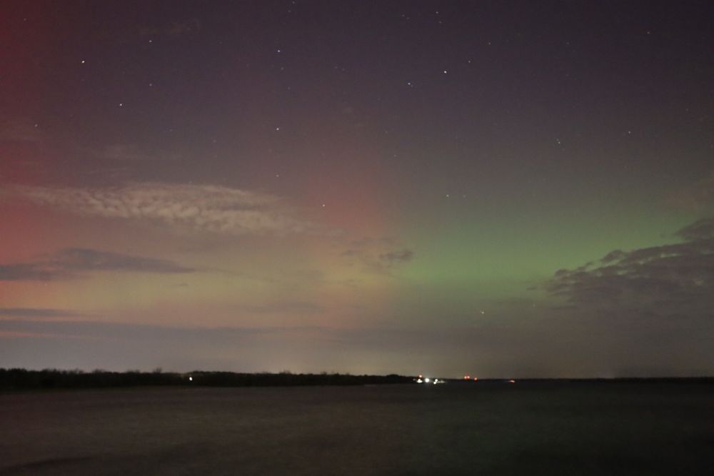Across a cold gray lake, the sky glows with a yellow-green near the horizon, partially blocked by low clouds. There are some vertical streaks of red forming, peeking up into the sky that is otherwise purplish and full of stars. 