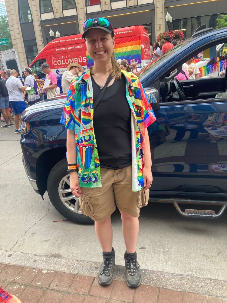 SK in hiking boots, cargo shorts, a black tank top and hat, and truly obnoxious rainbow button-up covered in cliche Pride symbols too large and loose so it’s worn like a cape. I’m standing in the queue for the Columbus pride parade