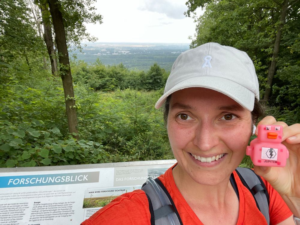 Author wearing running clothes with the pink duck in her hands, next to the sign “Forschungsblick”, in the background down in the valley is FZJ
