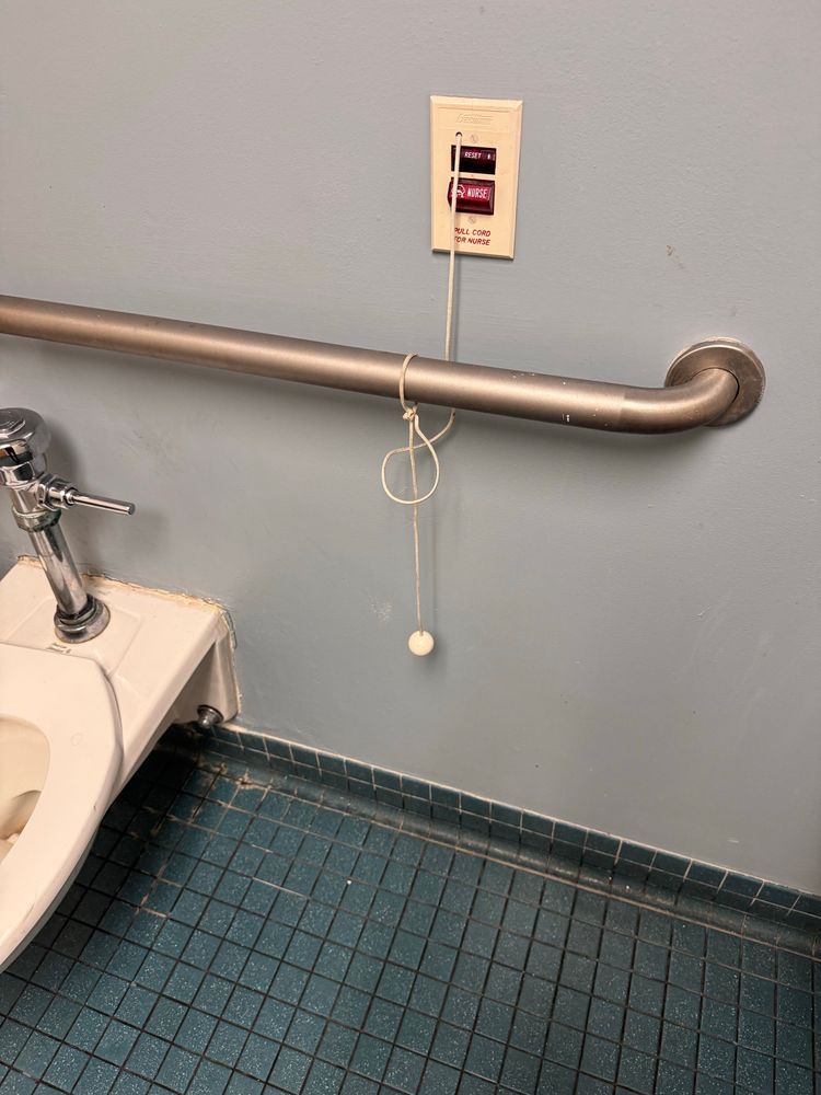 A public bathroom stall where a nurse call line is wrapped around and tied to a grab bar, lifting the tab several inches off the floor. 