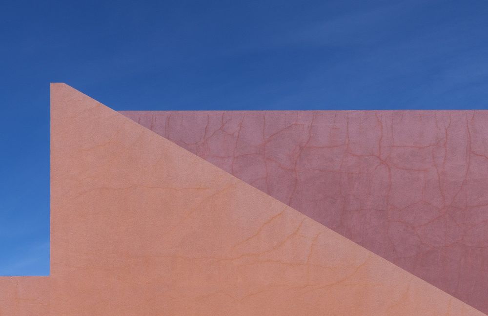 Minimalist photograph of an adobe wall. Its sunlit, coral-and-salmon surface is etched with dancing cracks. Geometry and shadow are in sharp play against a boundless blue sky.