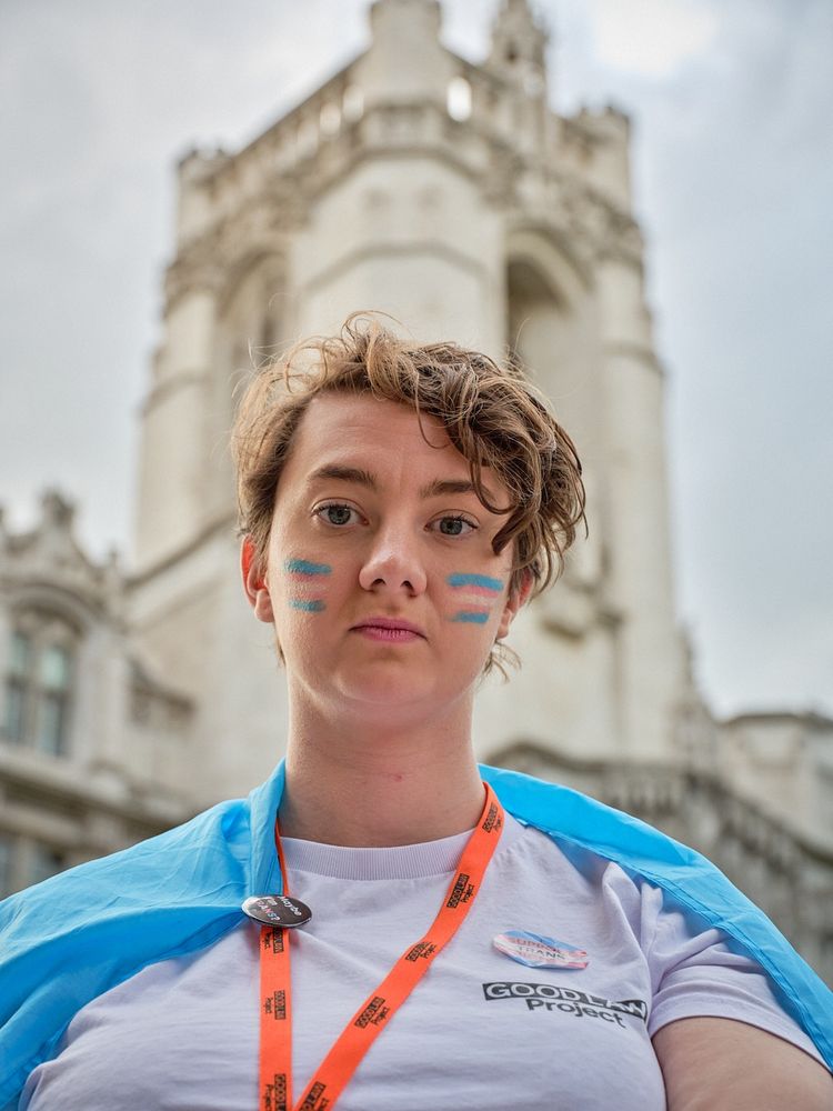 A close-up photo of me wrapped in a trans flag, wearing trans flag face paint. I am wearing a white Good Law Project tshirt and lanyard, with a Trans+ Solidarity Alliance badge which says Maybe I'm Trans? Behind me is the Supreme Court.
