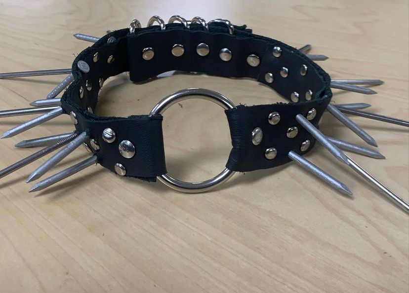 a photo of the hemorrhage collar design hand made by goliath. the black leather of the collar is dotted by rivets and long and short siding nails make up the spikes of the collar. a central medium sized o-ring holds both sides of the collar together as a unit. 