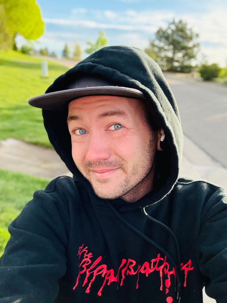 Selfie of Cortland walking outside. Bluesky and grass behind him. He’s wearing a black hoodie that says “The Blood Brothers” with the hood up. 