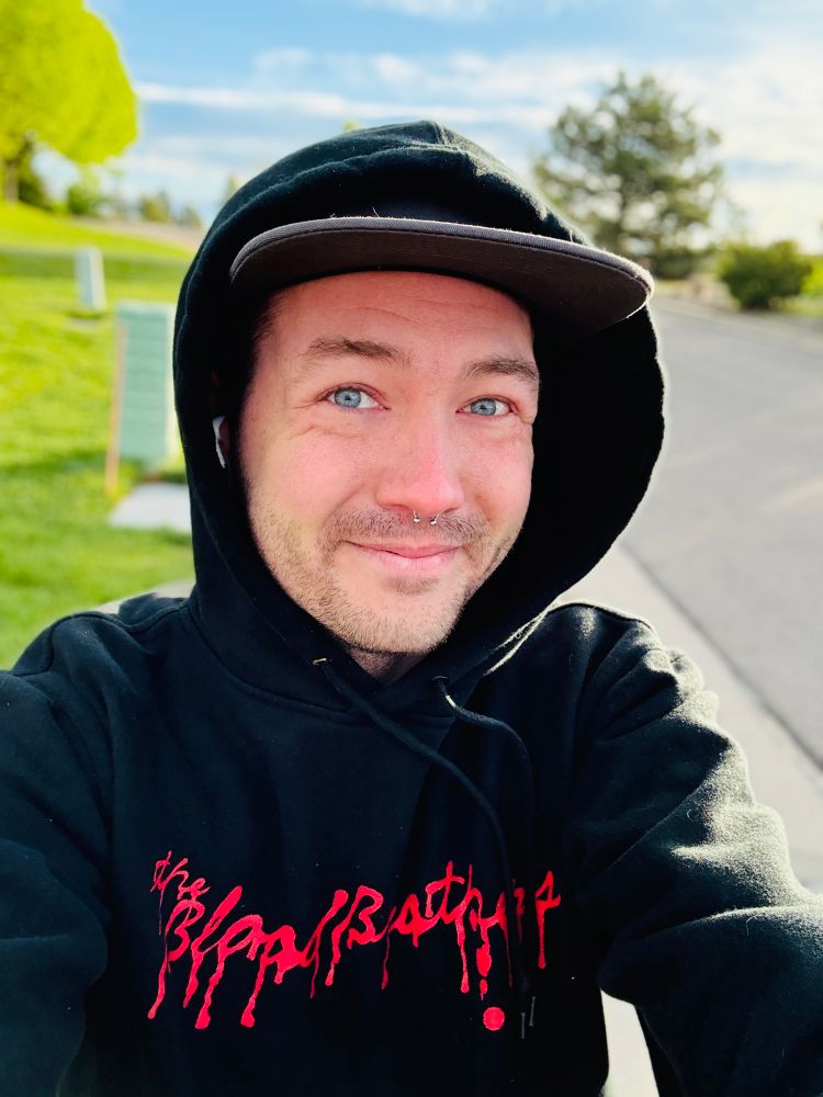 Selfie of Cortland walking outside. Bluesky and grass behind him. He’s wearing a black hoodie that says “The Blood Brothers” with the hood up. 