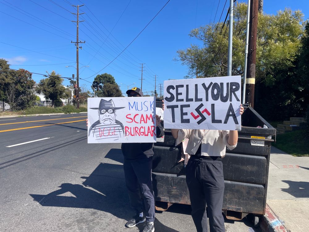 Two people holding signs against tesla and musk in in front of a dumpster on the side of the street