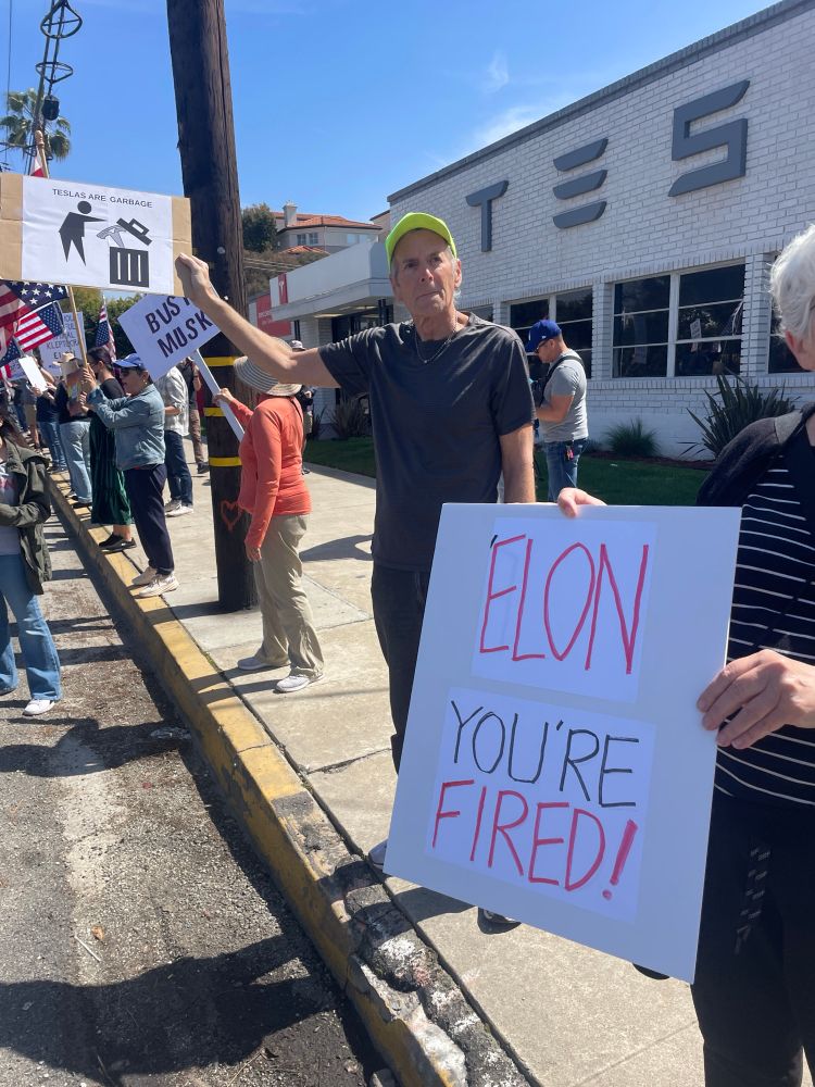 People on the sidewalk along the street holding signs against Tesla, Elon Musk, and Trump during the day
