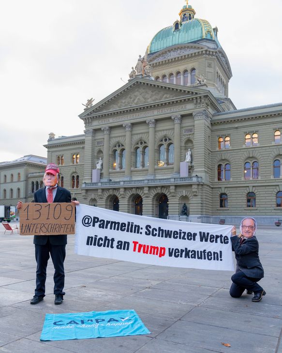 Vor dem Schweizer Bundeshaus stehen zwei Personen mit politischen Masken. Die Person links trägt einen dunklen Anzug und eine Gesichtsmaske von Trump. Sie hält ein Schild mit der Aufschrift „13'109 Unterschriften“. Die Person rechts kniet, trägt eine Gesichtsmaske von Bundesrat Parmelin und hält gemeinsam mit der anderen ein grosses Banner mit der Botschaft: „@Parmelin: Schweizer Werte nicht an Trump verkaufen!“ Auf dem Boden liegt eine Campax-Fahne. Das Bundeshaus bildet den Hintergrund der Szene.