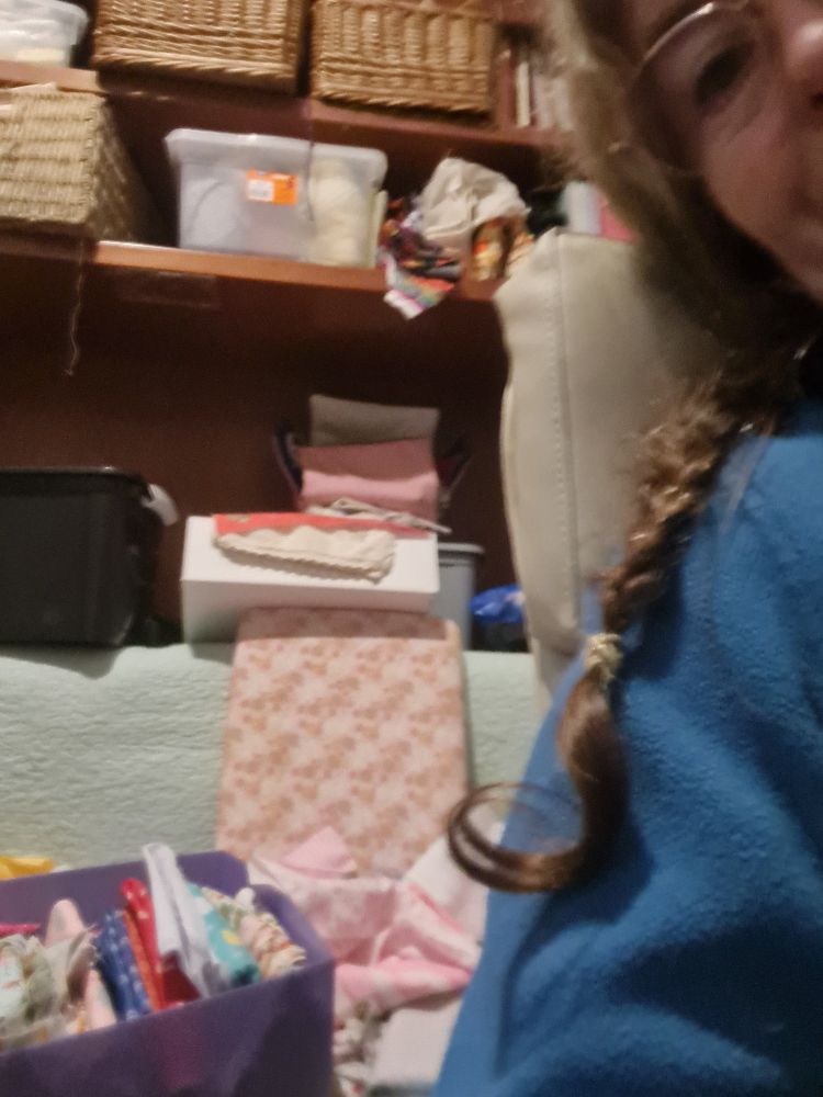 A direita mulher com óculos de trança e blusa azul. Atrás cesto de tecidos espalhados