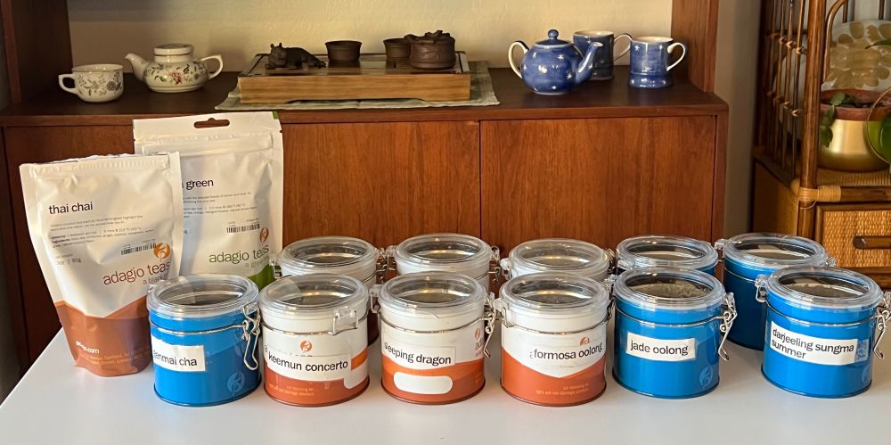 A collection of tea on a white table. There are two bags of loose leaf tea. The rest of the teas are in labelled tins, either blue or white and brown. In the background is a china cabinet with a display of tea ware sets.