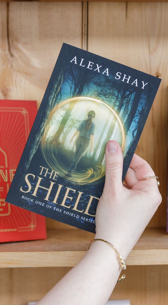 An arm coming up from the bottom holding a book, The Shield, in its hand. The Shield by Alexa Shay, Book One of the Shield Series. The cover is blue foresty scene with a girl walking into the woods with a gold circle around her.