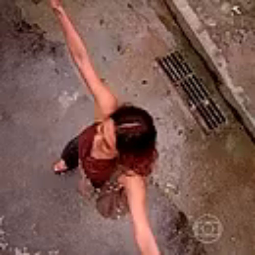 a woman is standing on a wet sidewalk with her arms outstretched and a drain in the background .