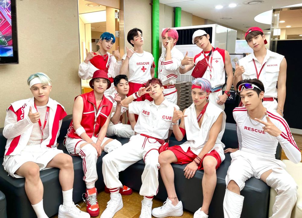 대기실에서 열한 명의 남자 아이돌이 구조대 콘셉트 의상을 입고 단체 포즈를 취하고 있다. 흰색과 빨간색이 포인트인 의상에 ‘RESCUE’ 글자가 새겨져 있으며, 각자 선글라스, 헤드밴드, 버킷햇 등 다양한 액세서리로 개성을 드러낸다. 모두 밝게 웃거나 손가락 하트를 하며 카메라를 향해 포즈를 취하고 있다. 에너지 넘치는 무대 대기 분위기다.