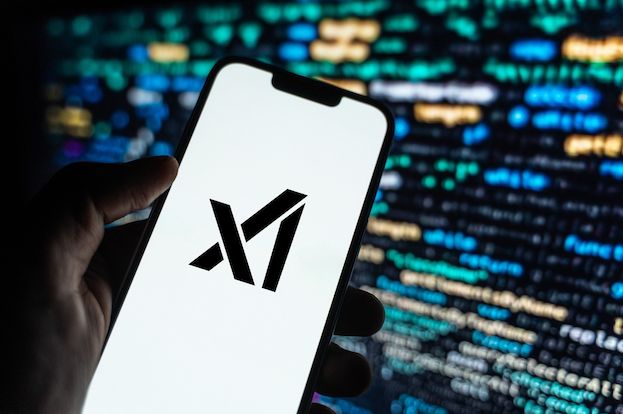 A photo of a hand holding a large phone with a white background and the xAI logo in black. Behind the phone is a lighted computer screen.