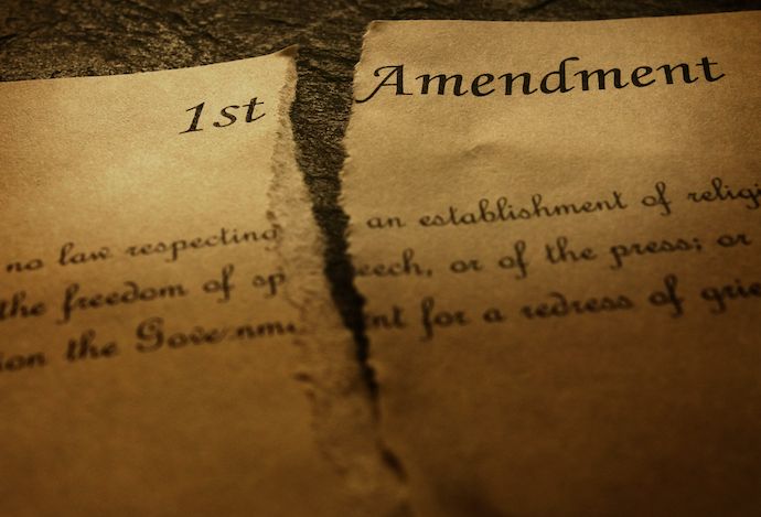 An image of a old, yellowed paper with the words 1st Amendment written on it and parts of the text from the amendment written in script. The paper is torn down the middle.