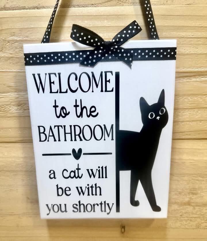 Door sign showing a black cat “welcome to the bathroom. A cat will be with you shortly”