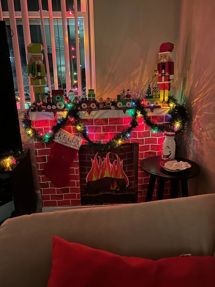 A cardboard fireplace with some lighted garland, two nutcrackers on top of it, a Christmas train that is actually stocking holders, and a stocking with my name written in glitter hung by the fire. 