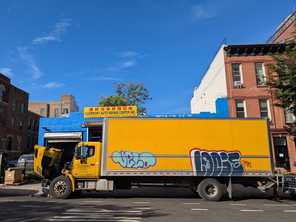 A yellow box truck with some graffiti, under a yellow sign with Chinese and English writing, under a blue sky, looking a bit all together like the Ukrainian flag