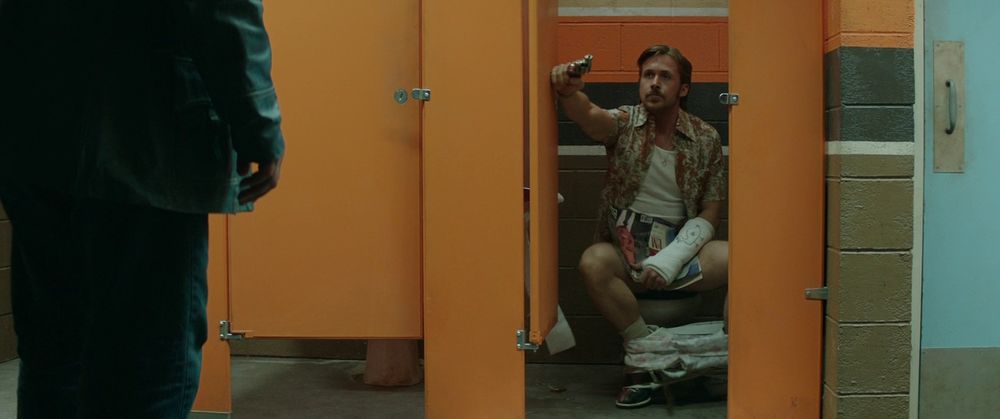 Image from the Nice Guys. Ryan Gosling is sitting on a toilet in a cubicle with his trousers around his ankles. He's trying to hold the door open with a gun in one hand.