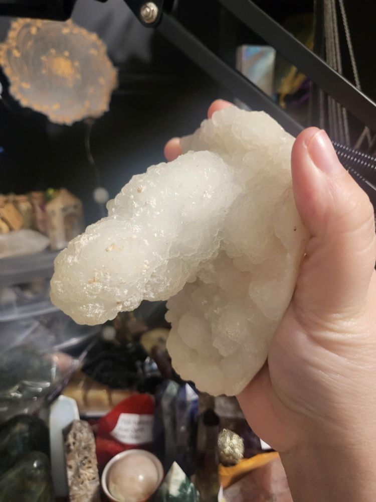 Kendra holds a white quartz stalactite resembling a penis at half-mast. In the background are a variety of crystals, a wooden wheel of the year magnet, and a phone mount.