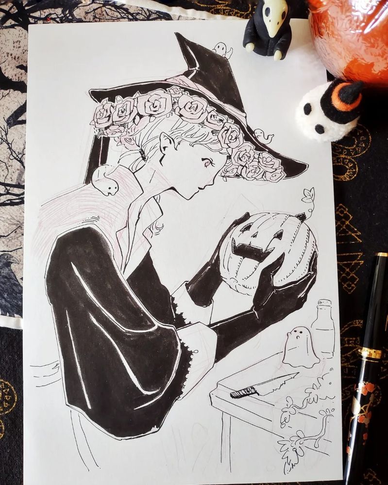 A top down photograph of a piece of paper.  The paper has a pen and ink illustration of a character holding a carved pumpkin.  The character is wearing a witch hat with roses around the brim and black gloves.  Placed around the paper are some Halloween themed miniatures and a fountain pen.