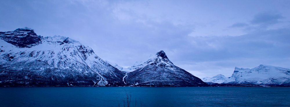 Blick auf eine Berglandschaft, in der Mitte ein Berg wie eine Zipfelmütze, im Vordergrund tiefblaues Wasser. Der Himmel ist bewölkt, aber es gibt helle Stellen. Die Berge sind schneebedeckt, aber man sieht auch schwarzes Gestein.