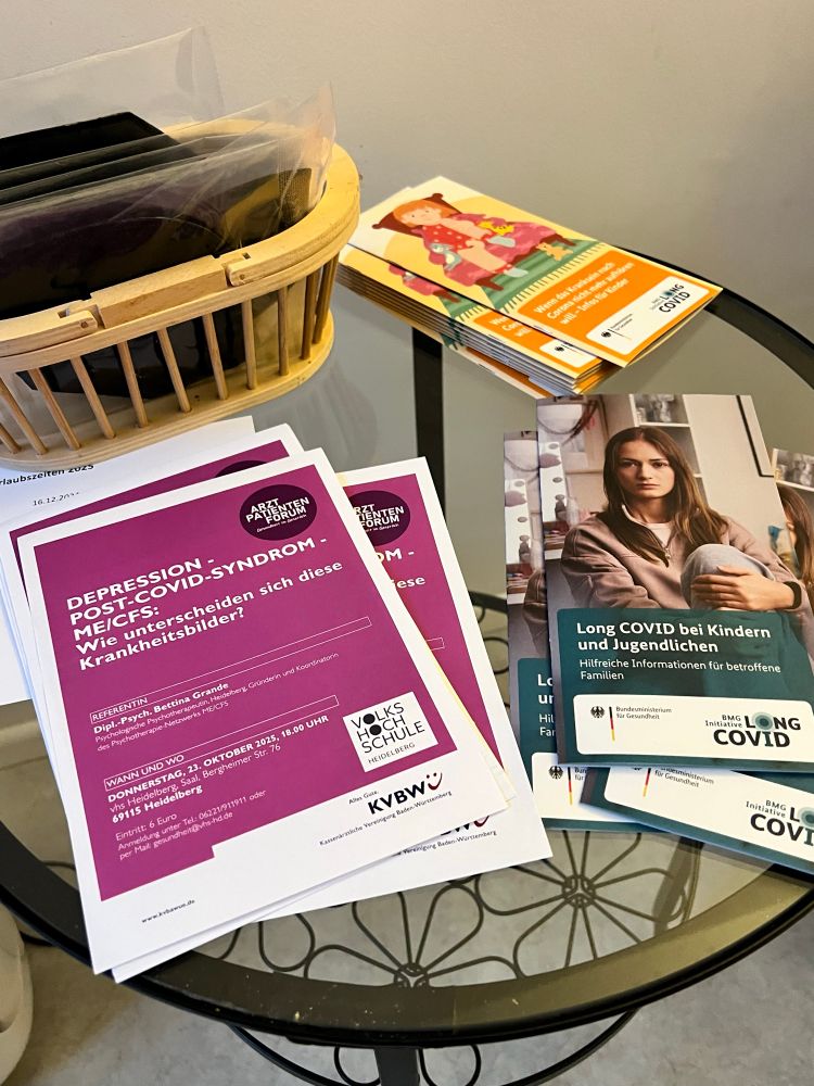 Ein kleiner Glastisch mit mehreren Infomaterialien. Links ein Stapel lilafarbener Plakate zur Veranstaltung „Depression – Post-COVID-Syndrom – ME/CFS“. Rechts Broschüren mit Titeln wie „Long COVID bei Kindern und Jugendlichen“ vom Bundesgesundheitsministerium. Im Hintergrund liegen weitere bunte Flyer, oben ein Korb mit FFP-2Masken
