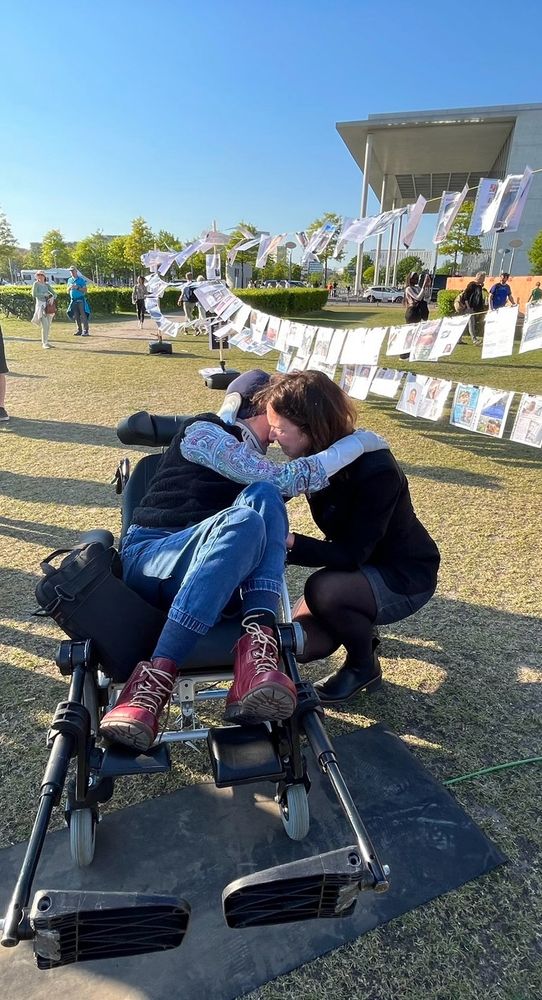 Umarmung zweier Frauen; eine liegt im Rollstuhl, die andere in der Hocke bei ihr