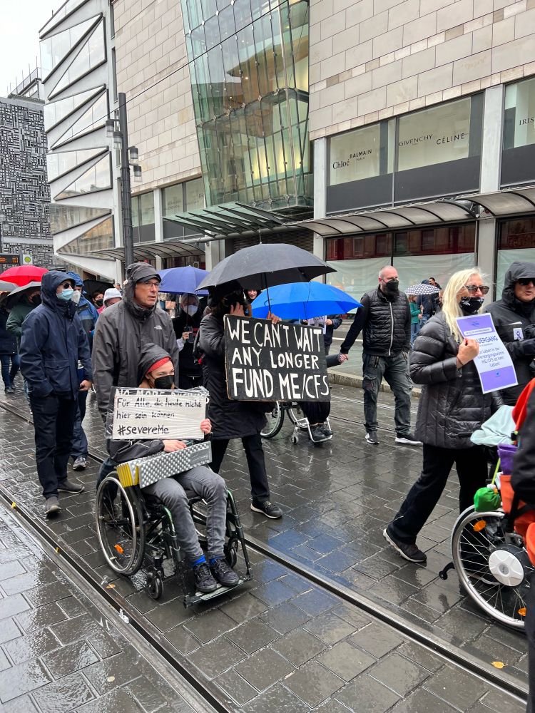 Demonstranten, davon 2 im Rollstuhl, Plakate für ME/CFS Awareness werden gezeigt.