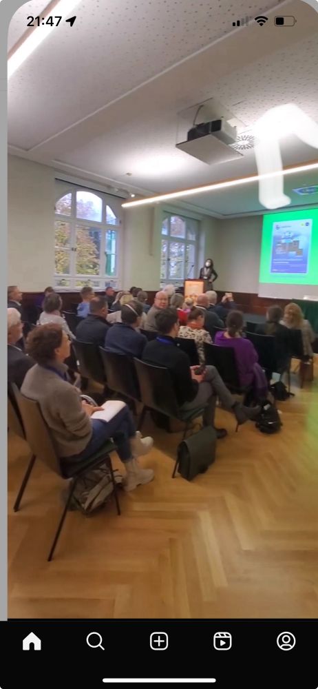 Vortragende (Bettina Grande) neben ihrer Präsentation, im Vordergrund das Publikum auf Stühlen sitzend.