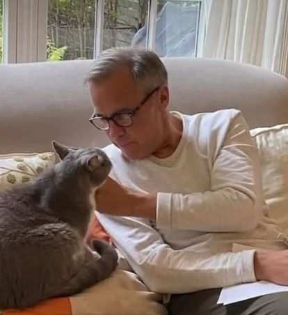 Prime Minister Mark Carney, dressed casually and wearing glasses, rubs the chin of his grey and white cat, Nico, while sitting on a couch. 