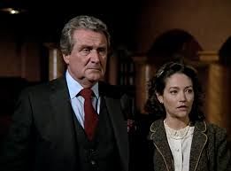 Grim-faced suited man stands next to dark-haired woman with shocked expression.