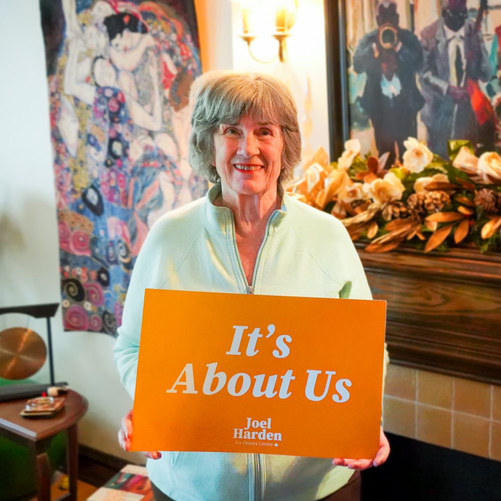 Diane holds a sign that says "It's about us" in white text on orange background