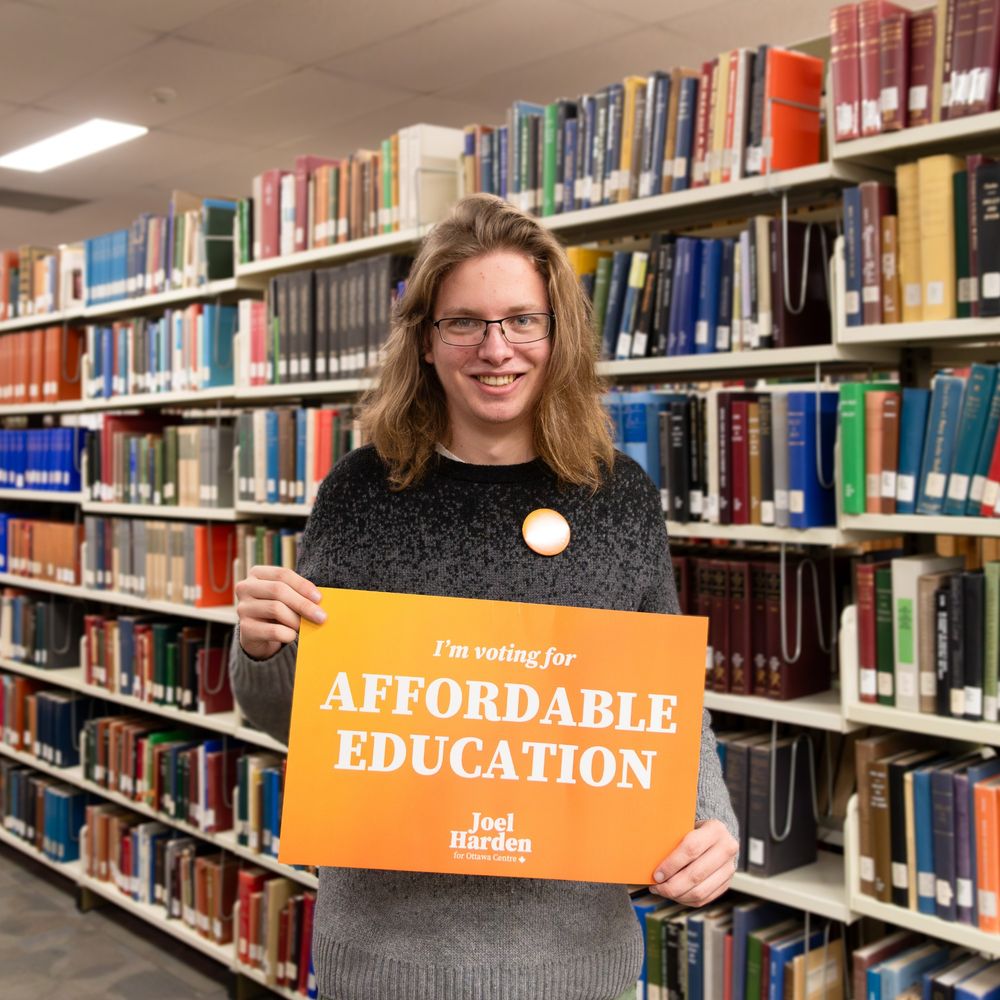 James stands next to a shelf of books in the Carleton University library. He is holding an orange sign that reads "I'm voting for Affordable Education"