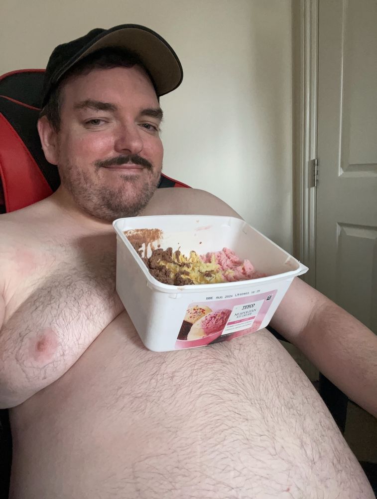 A fat gay dad looking type eating a 2litre tub of ice cream balancing it on his belly as if it's a table.