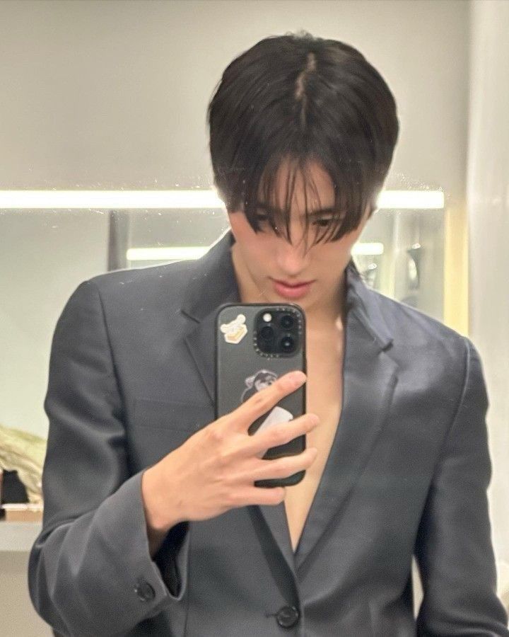 Minho segurando o celular em frente ao espelho usando um terno cinza.