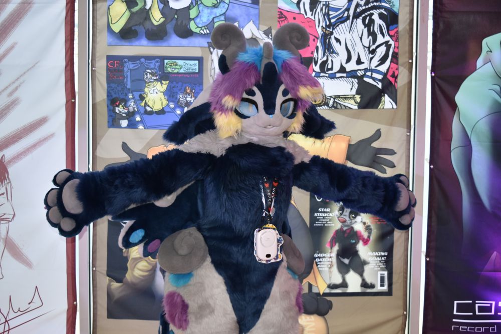 A photo of Parabella, a kemono character with dark blue and white fur, grey curled horns on her head and at her waist, wings, and blue/purple/yellow hair. She is standing with her arms stretched out, as if to gesture at her surroundings. She is standing in front of an illustration of Mel, a honey badger in a yellow jacket, who is doing the same pose.