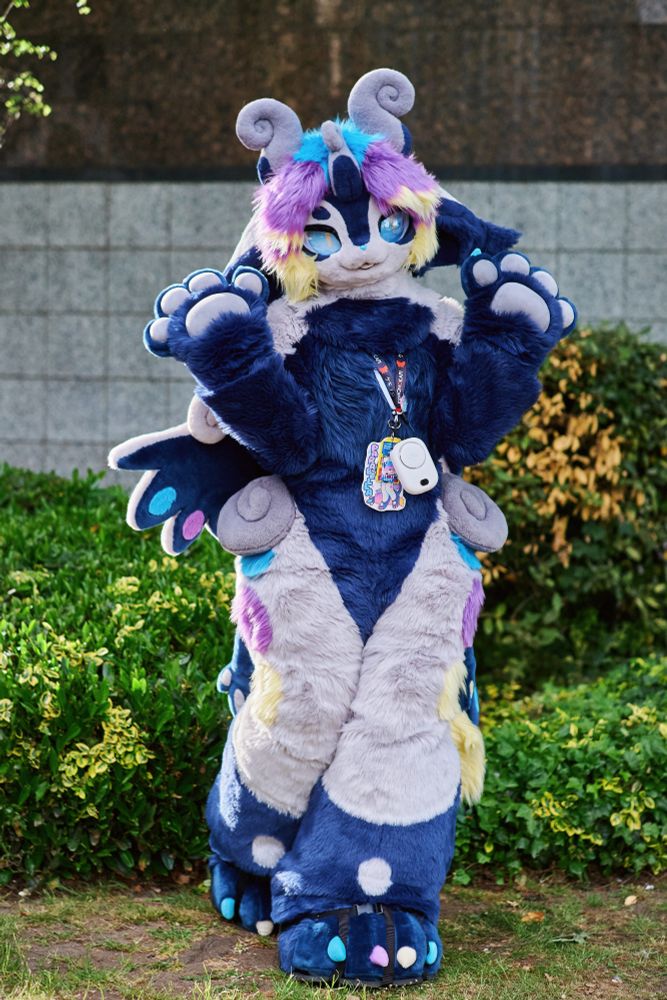 A photo of Parabella, a blue and white furred kemono creature with curled grey horns and blue/purple/yellow highlights. She is standing with both hands up and posing cutely for the camera, with some green bushes behind her.