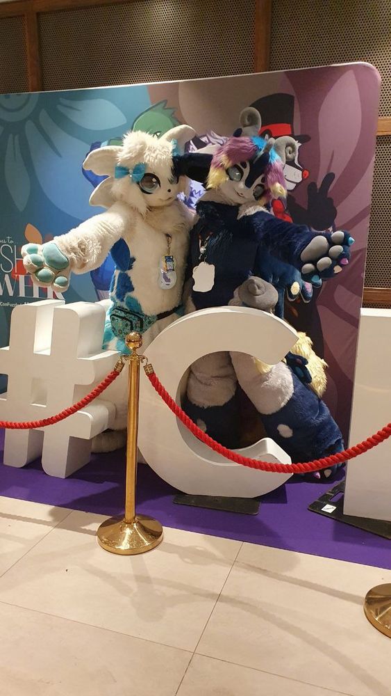 Numazu and Parabella standing behind big standee letters reading # and C, behind a red velvet rope. They are holding their hands outstretched towards the camera.