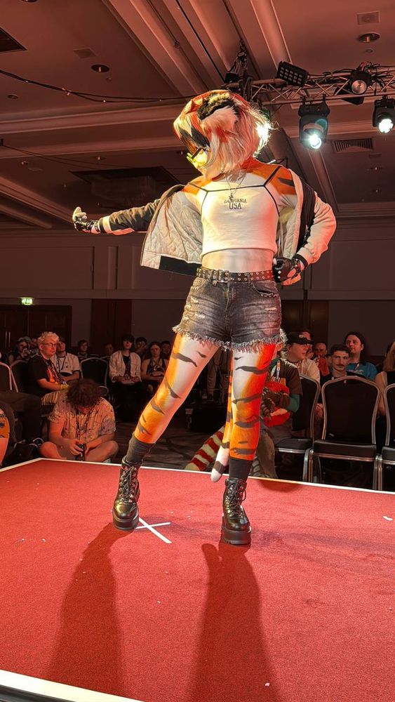 A photo of Waai Fu, an anthromorphic tiger girl from the videogame Arknights, standing on a red catwalk in profile. She is facing the left of the image with her chest towards the camera, holding out her right hand and gesturing in a "come here" motion, while her left arm is on her hip. She is wearing a bomber jacket, a white sleeveless crop top, and ripped denim shorts.