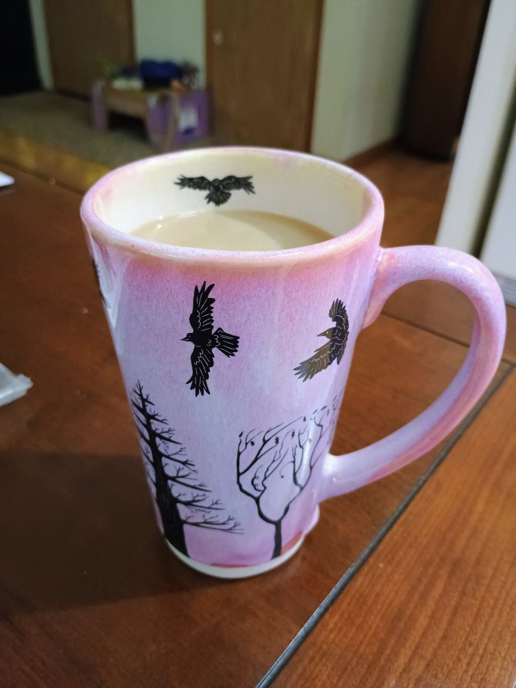 A large pink mug with black trees and crows on it from @surlyamy.bsky.social filled with a vanilla latte sitting on a cherry wood table