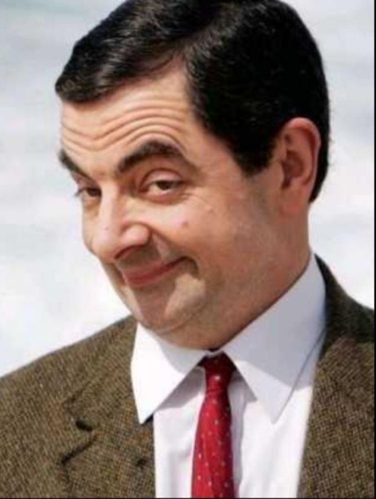 Rowan Atkinson as MR bean, wearing a white shirt, brown suit jacket and red tie. He has a smug look on his face 