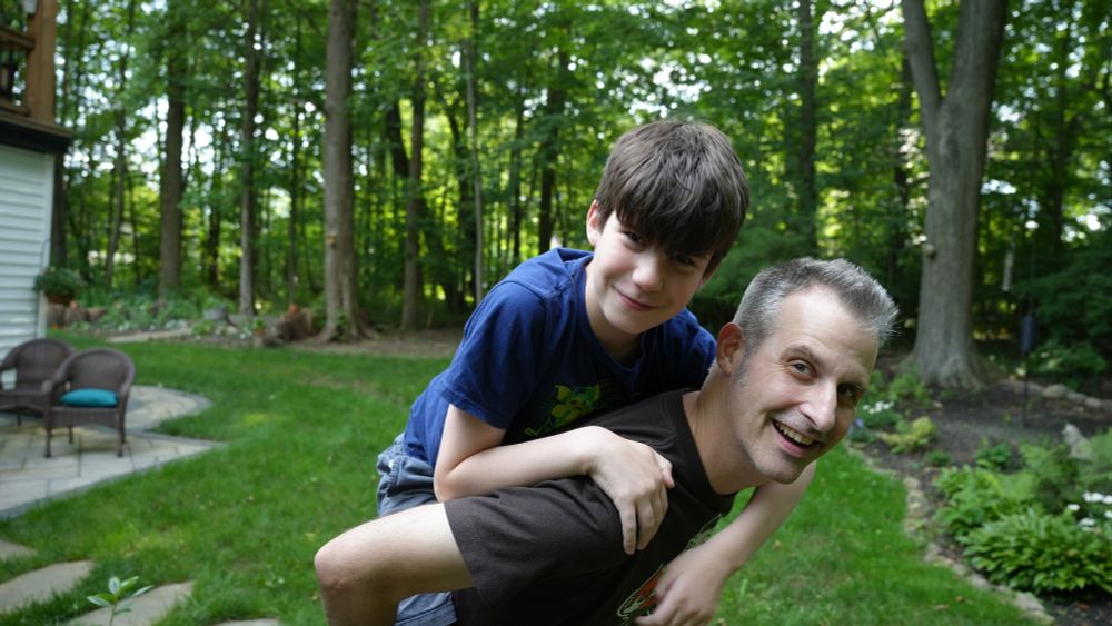 A man (Nathaniel Bowler) is standing in the grass and carrying a child (Santiago Hidalgo-Bowler) on his back. Santiago has his hands over Nate’s shoulders. They are both smiling and there are trees behind them. They are outside and it is daytime.