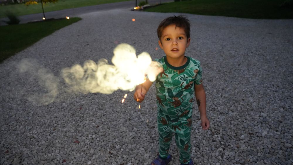 A child stands and holds a sparkler facing up on a driveway. He is outside and it is nighttime.