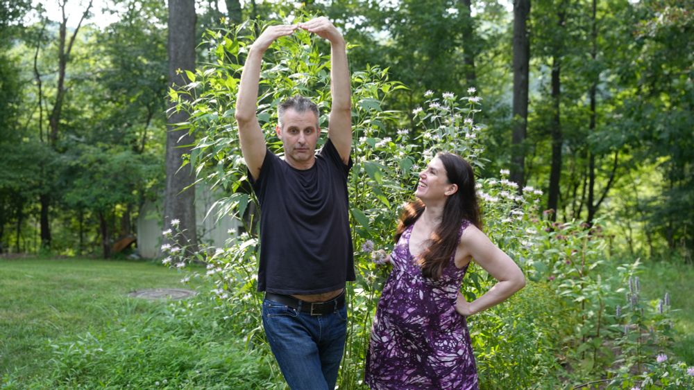 A man (Nathaniel Bowler) and a woman (Alexandra Hidalgo) stand and pose in front of a native garden. Nate is posing like a ballerina and Alex is looking up at him with a smile. There are trees behind them. They are outside and it is daytime.