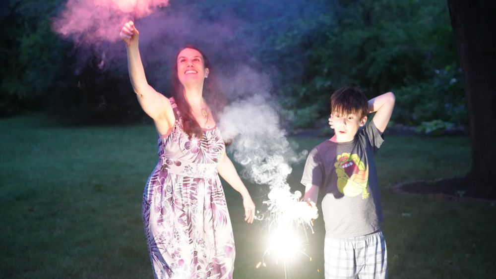A woman, Alexandra Hidalgo, is holding a sparkler up above her head, and she is looking up at it. To her right, a child, Santiago Hidalgo Bowler, holds a sparkler out in front of him that is a bright white, illuminating his face and body. He looks at the sparkler with his eyes wide and mouth agape. They are both outside, with grass and trees behind them. It is nighttime.