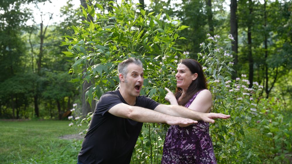 A man (Nathaniel Bowler) and a woman (Alexandra Hidalgo) stand and pose in front of a native garden. Nate has his mouth open in a shocked expression, his hands outstretched in front of him Alex is smiling at him and pointing behind them. There are trees behind them. They are outside and it is daytime.