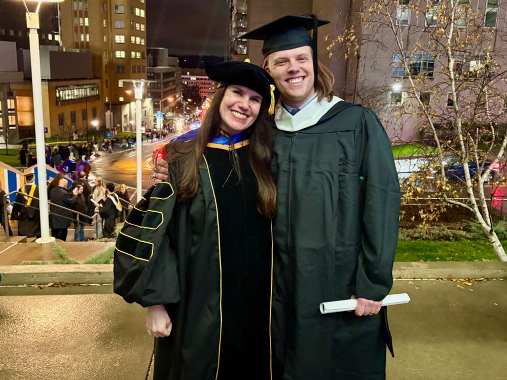 A woman in professor regalia (big sleeves in her robe, fluffy velvet hat) stands with a man in traditional graduate cap and gown. He is holding a rolled up diploma. They both smile at the camera. Behind them there are buildings and winter trees. Other graduates can be seen in the distance. It is night time and it has been raining so the pavement is shiny. 